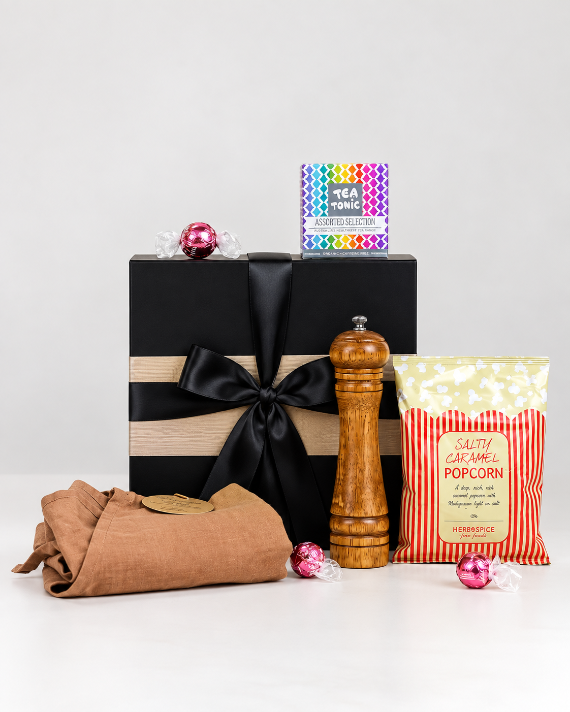 Home Sweet Home Housewarming Gift Box NZ featuring a wooden pepper grinder, linen apron, Tea Tonic favourites tea selection, Herb & Spice Fine Foods caramel popcorn, Lindt Strawberries & Cream Lindor chocolates and personalised card in a premium black magnetic gift box.