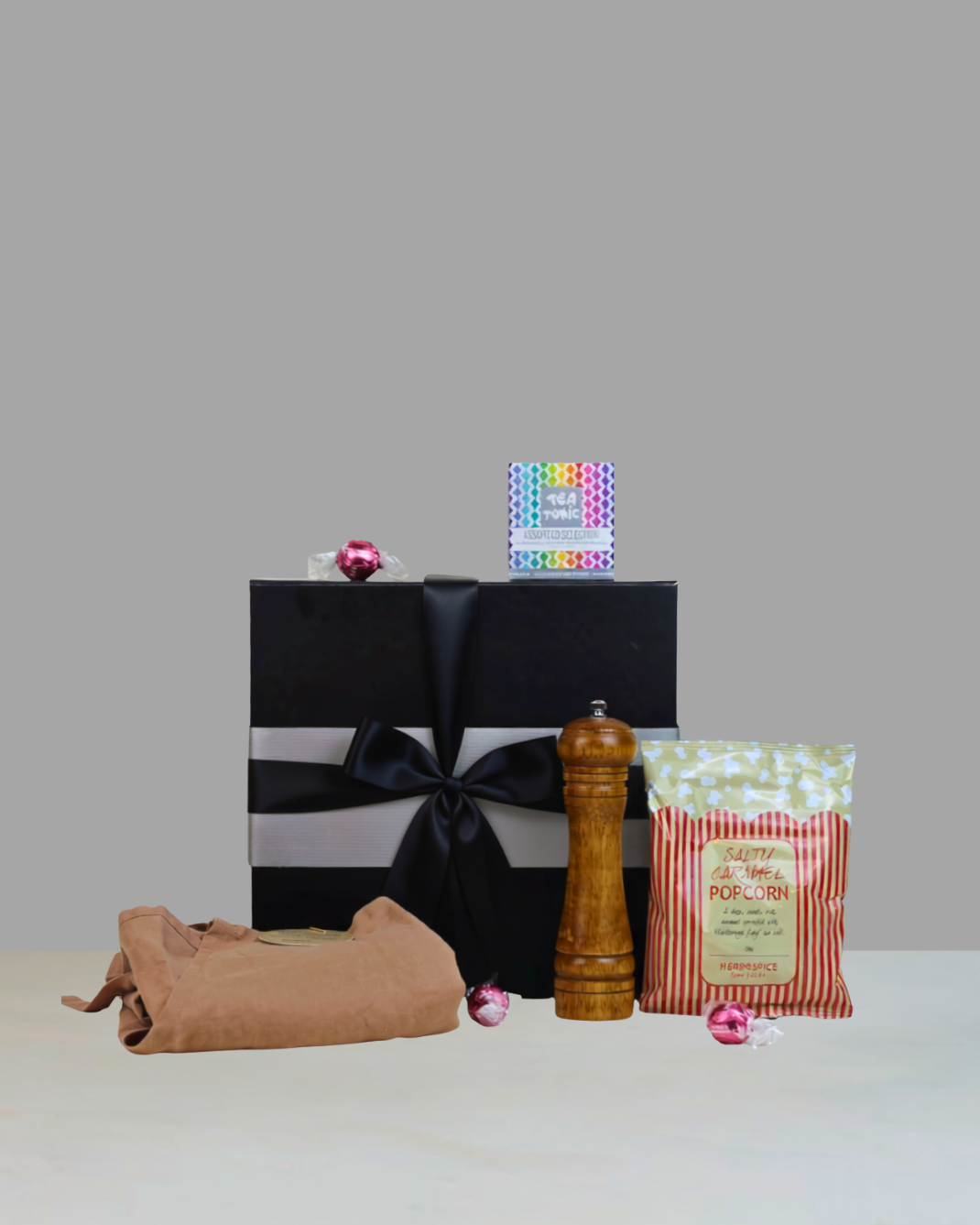 Home Sweet Home Housewarming Gift Box NZ featuring a wooden pepper grinder, linen apron, Tea Tonic favourites tea selection, Herb & Spice Fine Foods caramel popcorn, Lindt Strawberries & Cream Lindor chocolates and personalised card in a premium black magnetic gift box.