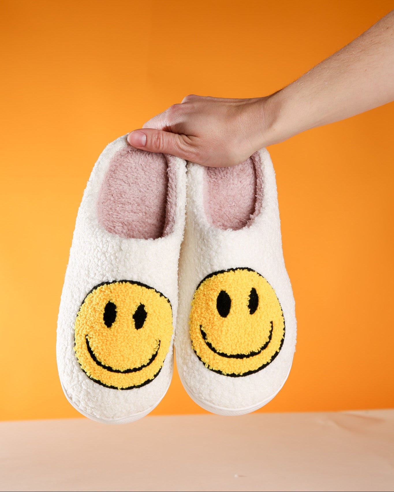 White slippers with smiley face design held by a hand against an orange background
